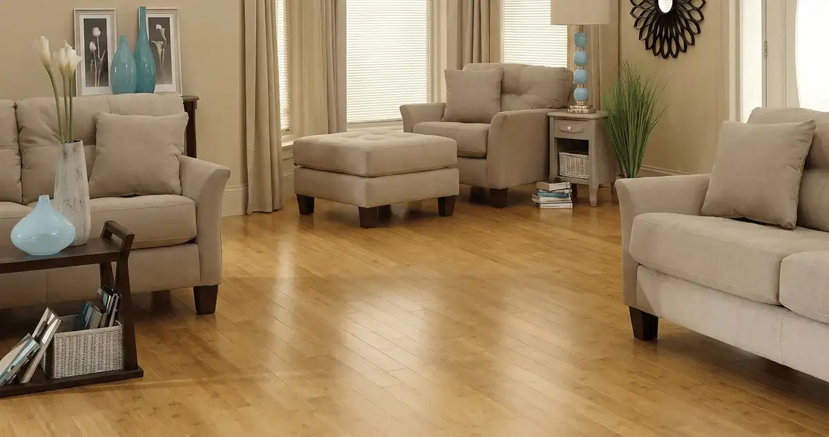 A living room with professionally installed bamboo flooring, beige sofas, a matching ottoman, a side table with a blue lamp, vases, framed pictures, and large windows with blinds and curtains in Lakewood, CO